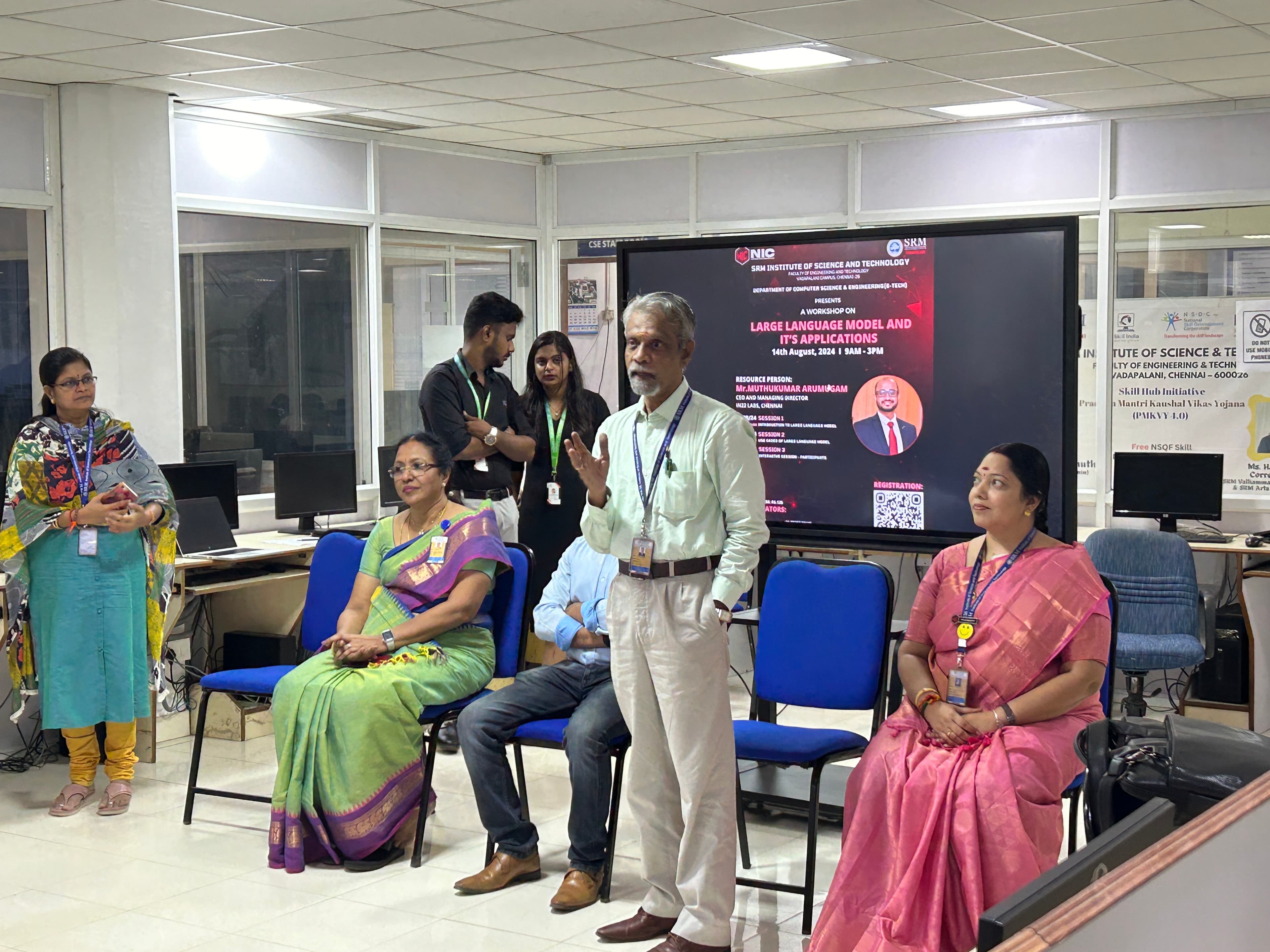 The Large Language Models workshop in a computer lab, faculty seated and standing in front of a wall screen carrying the event banner, one speaker addressing the room.