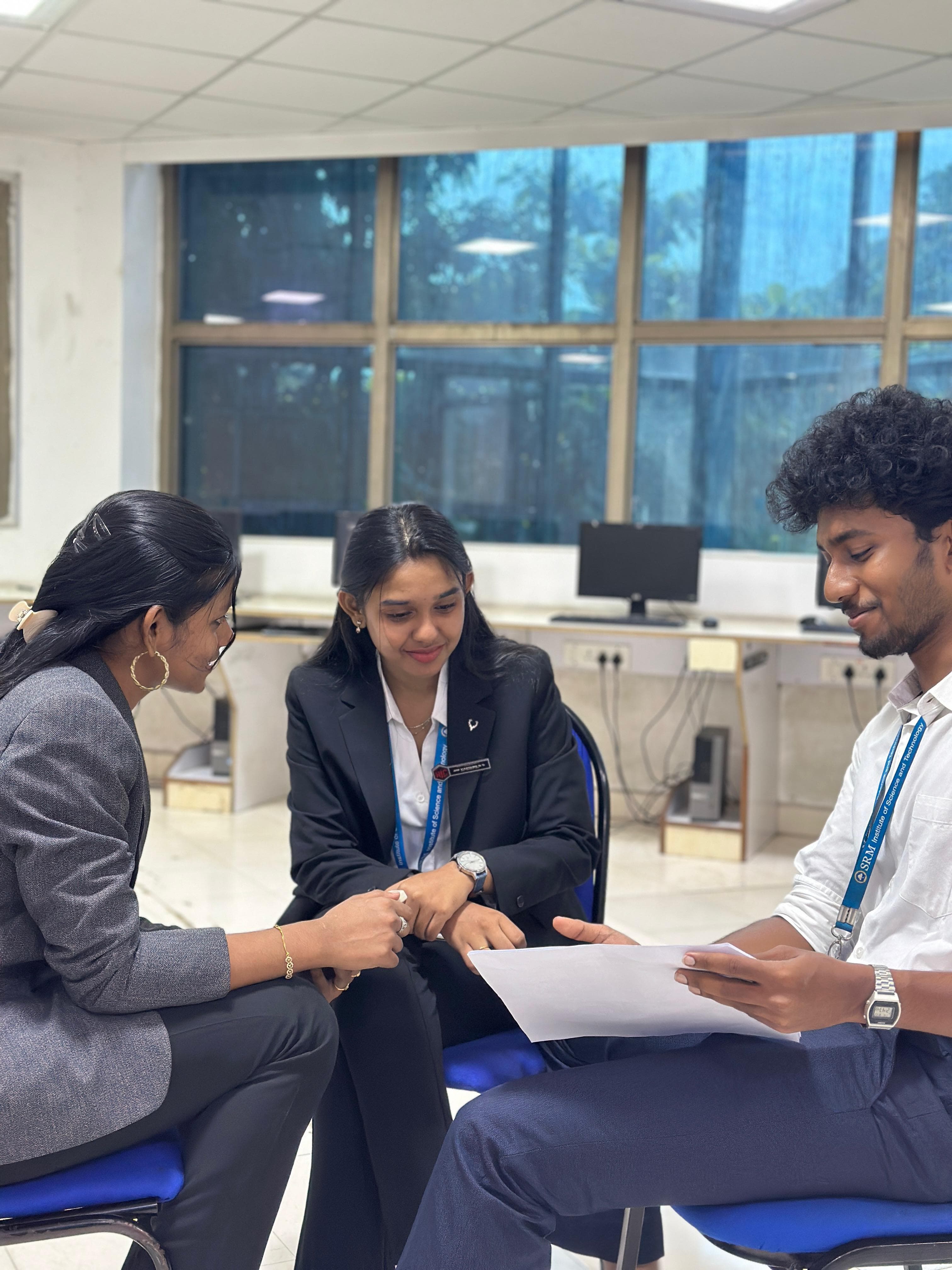 Three students in formal blazers and lanyards sitting together in a computer lab, reading over a printed sheet.