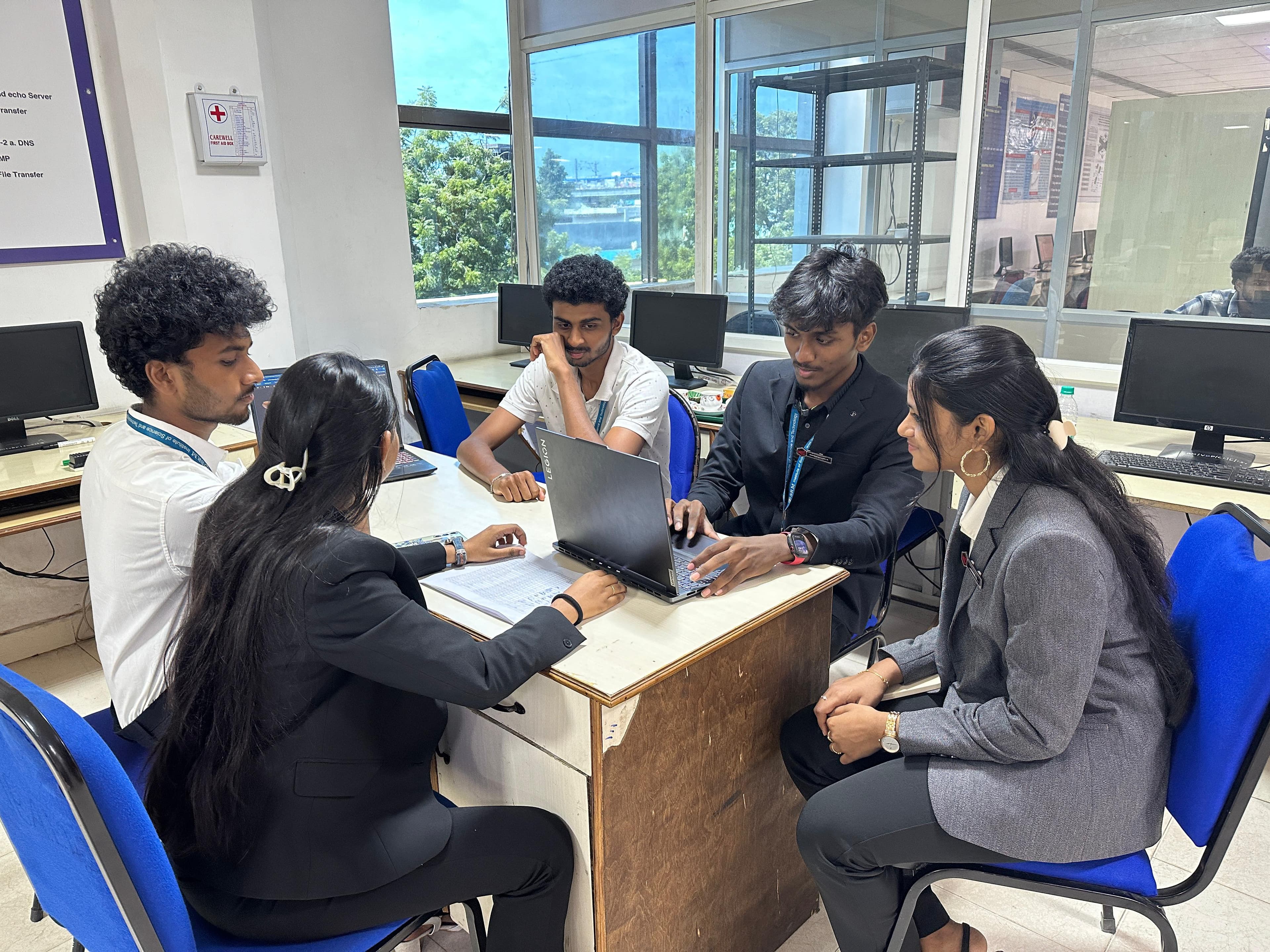 Five students around a lab desk with a laptop open between them and a notebook laid out beside it, working through something together.